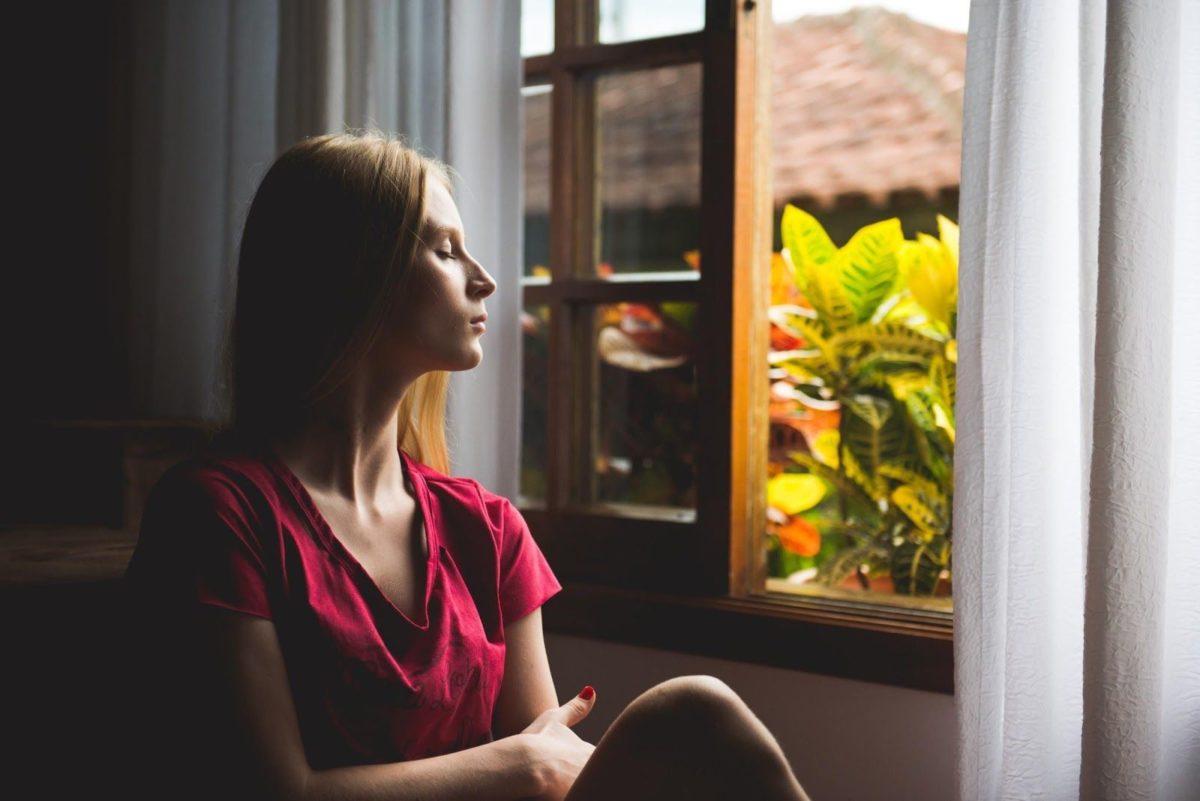 Young woman siting next to open window with eyes closed
