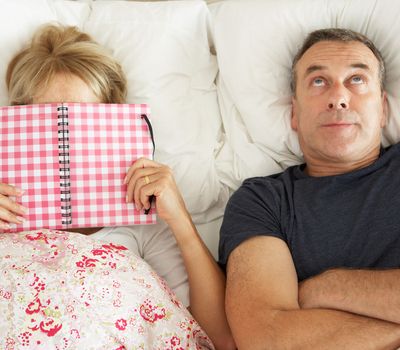 Bored Looking Senior Man Lying In Bed Next To Senior Woman Reading Book