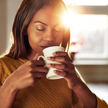 Attractive young black woman sitting at a table at home backliot by a bright sun enjoying a cup of fresh coffee with her eyes closed in bliss