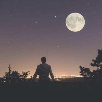 Young man sitting on the wall at night. City lights, moon and stars in the background