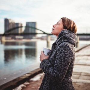 A young woman standing close to river with her eyes closed, holding cup of coffee, face toward the sun, contemplating the moment.