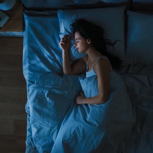 Top View of Beautiful Young Woman Sleeping Cozily on a Bed in His Bedroom at Night. Blue Nightly Colors with Cold Weak Lamppost Light Shining Through the Window.